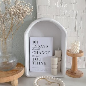 May include: White ceramic arched shelf with a white book titled "101 Essays that will Change the way You Think" by Brianna Wiest. The shelf is on a white textured wall with the text "Maya & Havana Art Studio". There are three white candles on a wooden stand to the right of the shelf. The shelf is 25 cm wide, 35 cm high, and 13 cm deep.