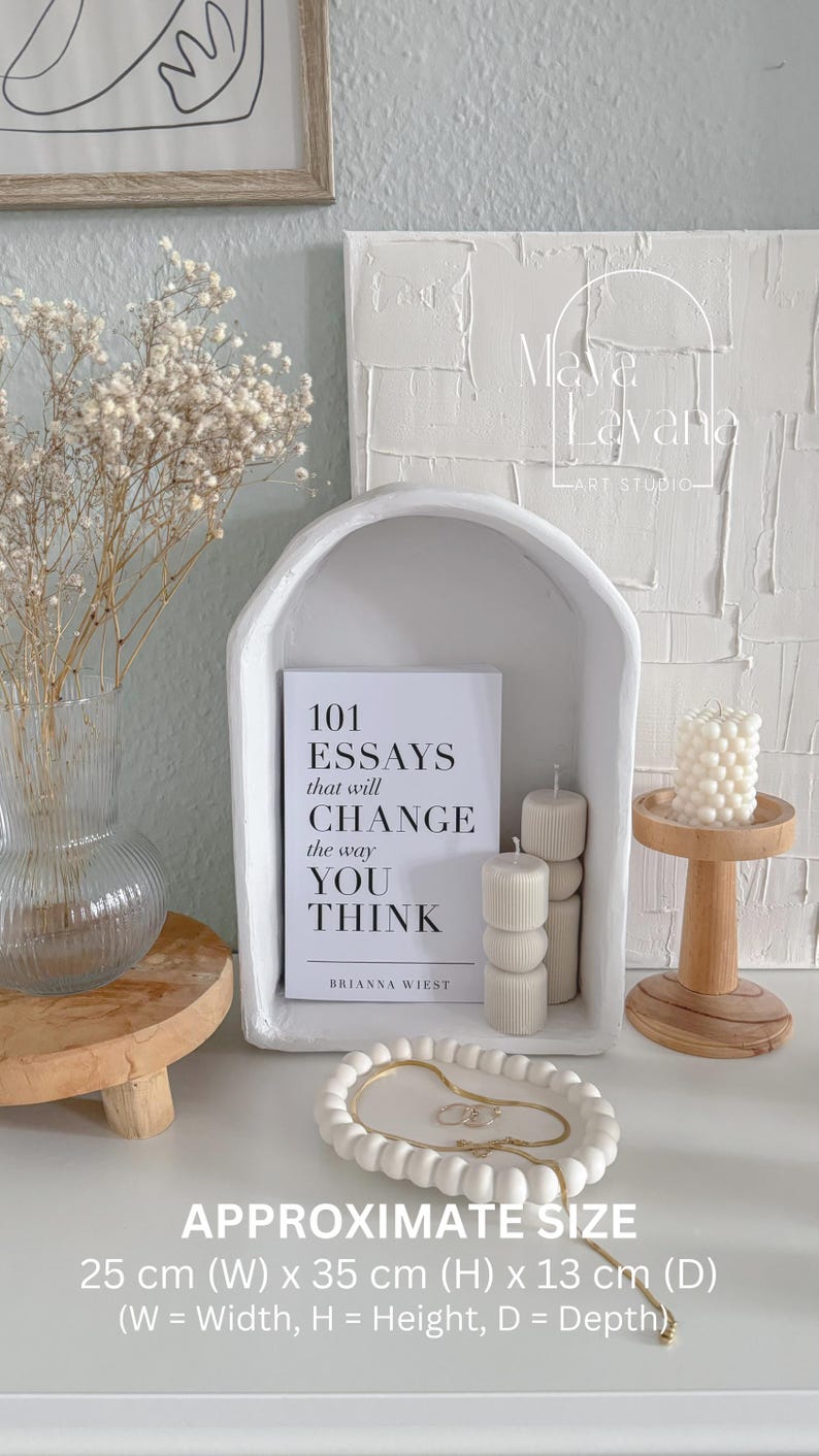 May include: White ceramic arched shelf with a white book titled "101 Essays that will Change the way You Think" by Brianna Wiest. The shelf is on a white textured wall with the text "Maya & Havana Art Studio". There are three white candles on a wooden stand to the right of the shelf. The shelf is 25 cm wide, 35 cm high, and 13 cm deep.