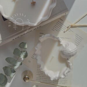May include: Two white ceramic candle holders shaped like seashells. The candles are white and have wooden wicks. The candles are surrounded by eucalyptus leaves and a gold candle snuffer.
