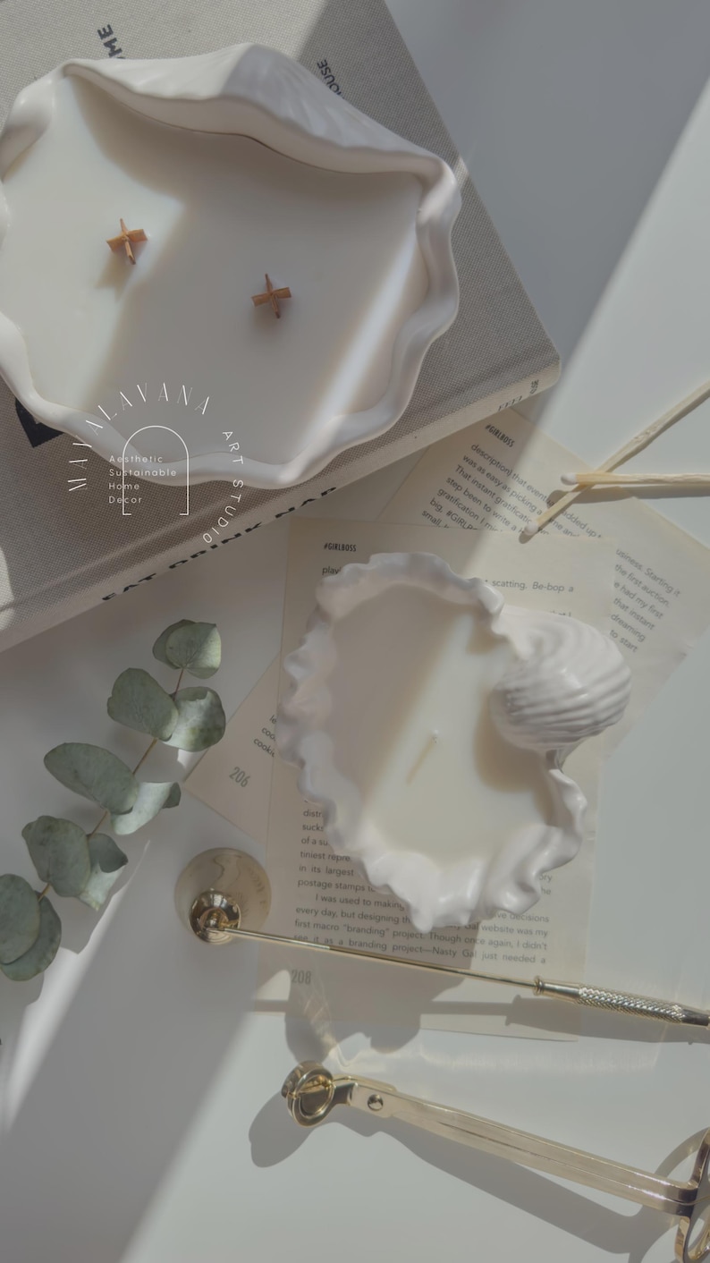 May include: Two white ceramic candle holders shaped like seashells. The candles are white and have wooden wicks. The candles are surrounded by eucalyptus leaves and a gold candle snuffer.