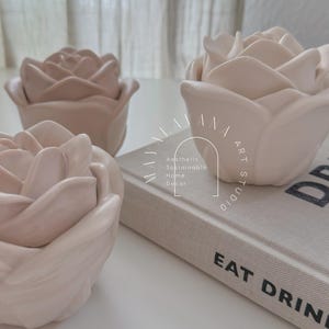 May include: Three pale pink rose-shaped ceramic containers on a white surface. The containers are arranged in a row, with the middle container resting on a book with the text "Eat Drink" visible. The book also has a logo that reads "Aesthetic Sustainable Home Decor."