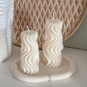 May include: Two white, wavy, cylindrical candles on a white ceramic tray. The candles are arranged side by side.