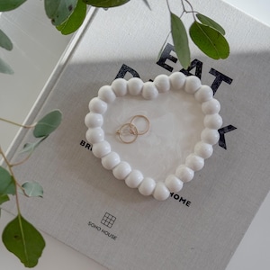 May include: White ceramic heart-shaped trinket dish with a beaded edge. The dish is sitting on a grey book with the text "Eat Drink Home" and "SOHO HOUSE" printed on it.