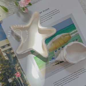 Może przedstawiać: Białe dekoracyjne naczynie w kształcie rozgwiazdy i białe naczynie w kształcie muszli spoczywają na magazynie. Magazyn przedstawia scenę na plaży z żółtym parasolem. Widoczny jest tekst "OF PLACE".