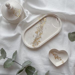 May include: A set of three decorative items: a lidded jar, an oval tray, and a heart-shaped dish. All are off-white with gold leaf accents. The items are arranged on a white, textured surface.