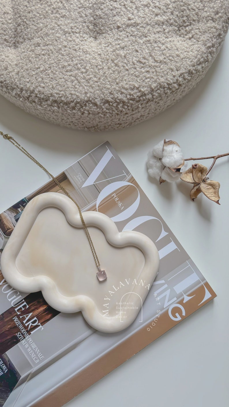 May include: A white ceramic tray shaped like a bear with a gold chain necklace with a pink stone pendant. The tray is on top of a magazine with the text "Vogue Art" and "POLSKA" visible. The background is a white surface with a white fluffy object and a dried cotton branch.