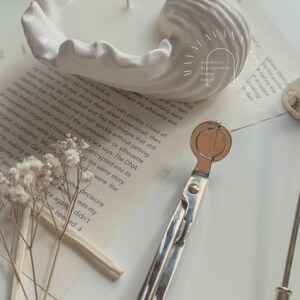 May include: A white ceramic candle holder shaped like a seashell with a white candle inside. The candle holder is sitting on a book with the text "Aesthetic Sustainable Home Decor" visible. A silver candle snuffer is also in the image.