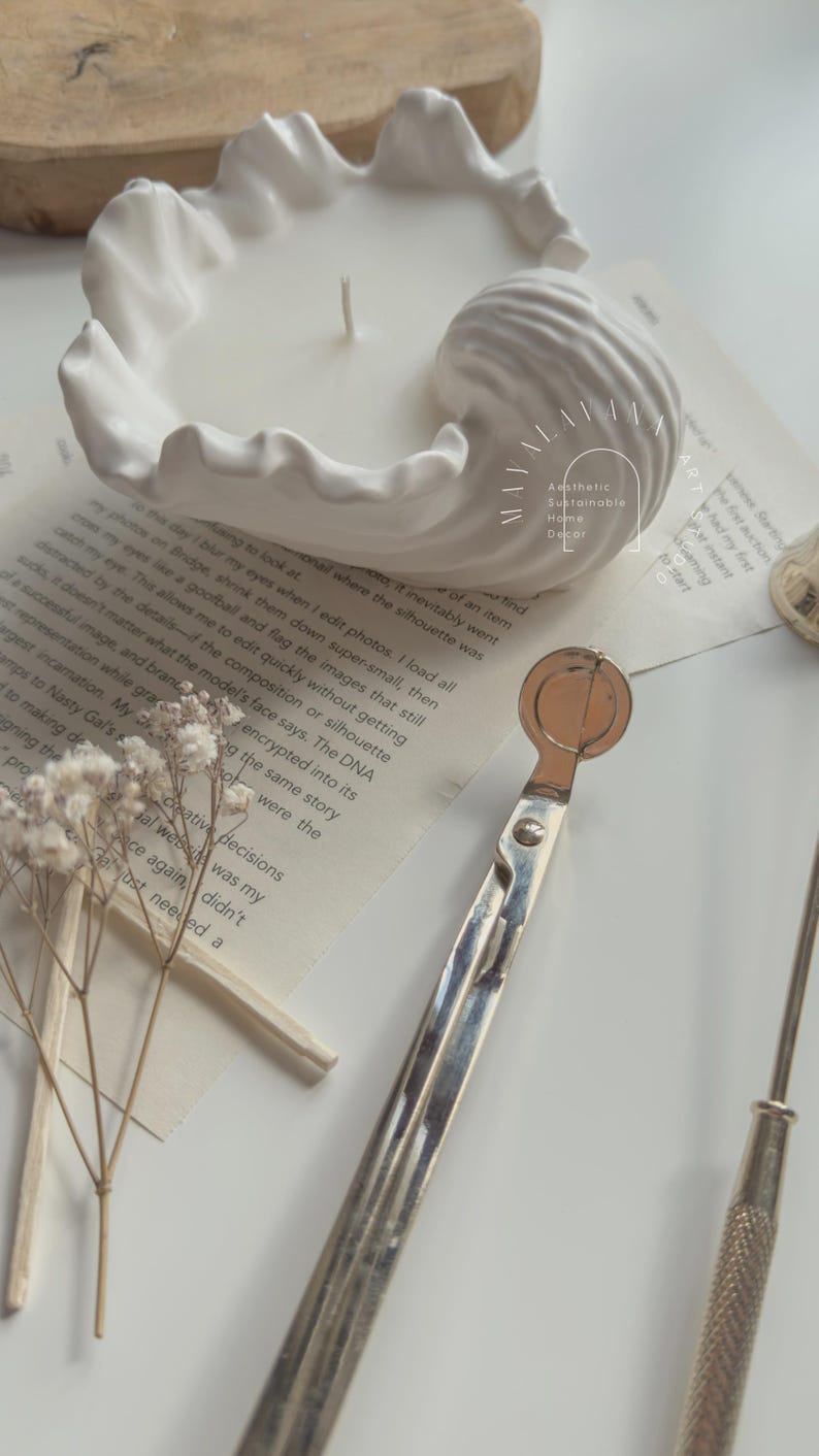 May include: A white ceramic candle holder shaped like a seashell with a white candle inside. The candle holder is sitting on a book with the text "Aesthetic Sustainable Home Decor" visible. A silver candle snuffer is also in the image.