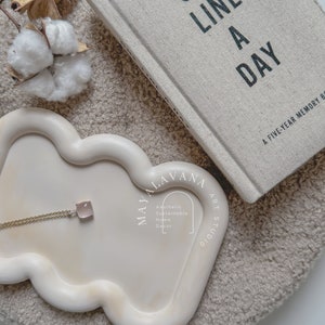 May include: A white ceramic tray with a wavy edge, featuring a gold chain necklace with a pink stone pendant. The tray is on a beige fluffy surface, with a book titled "One Line A Day: A Five-Year Memory Book" in the background.