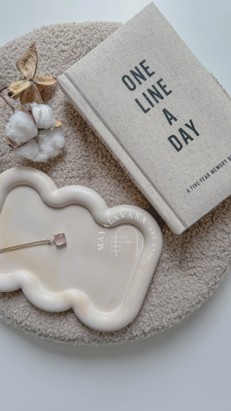 May include: A white ceramic tray with a wavy edge, featuring a gold chain necklace with a pink stone pendant. The tray is on a beige fluffy surface, with a book titled "One Line A Day: A Five-Year Memory Book" in the background.