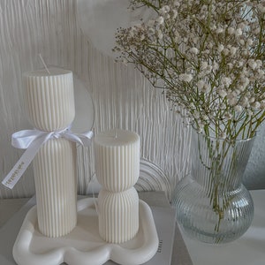 May include: Two white ribbed candles on a white cloud-shaped tray. The candles are on a book with the text "Los Angeles" and "Cereal" visible. A bouquet of white baby's breath flowers in a clear glass vase is in the background.