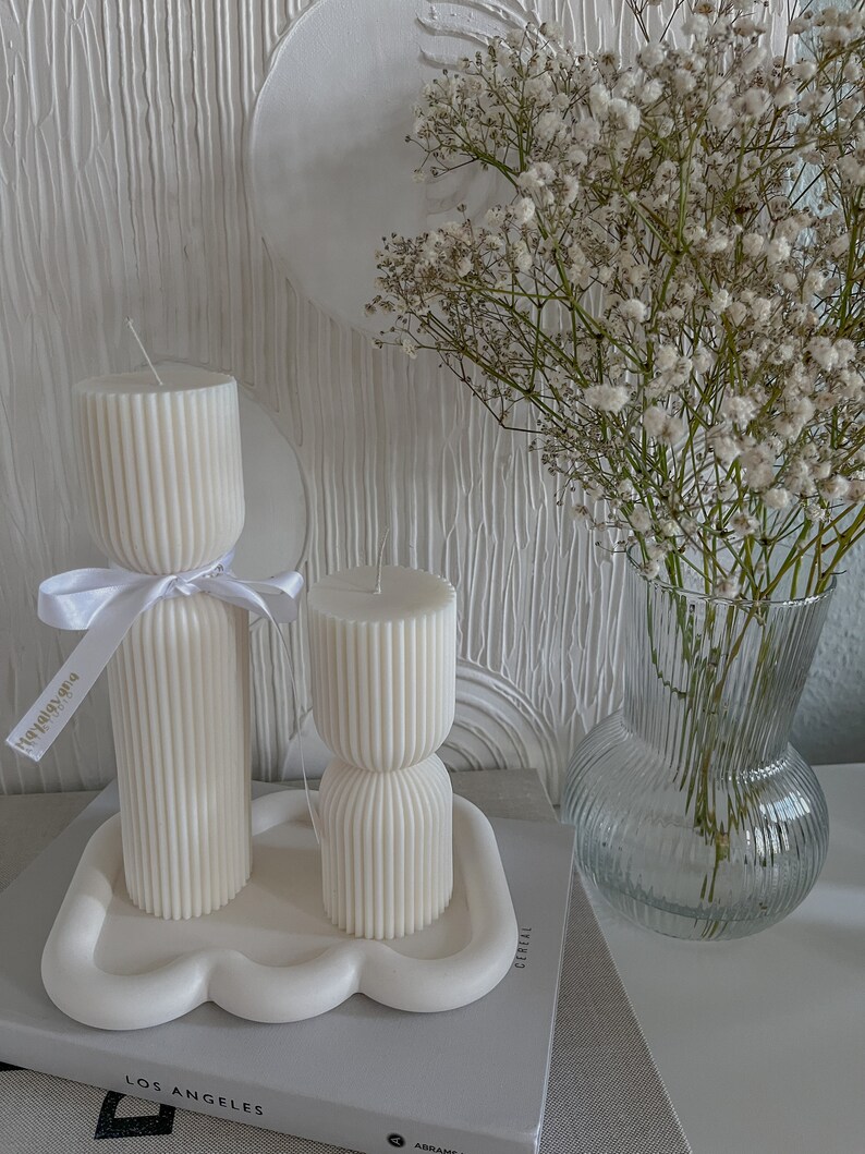 May include: Two white ribbed candles on a white cloud-shaped tray. The candles are on a book with the text "Los Angeles" and "Cereal" visible. A bouquet of white baby's breath flowers in a clear glass vase is in the background.