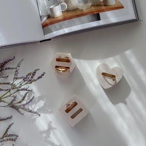 Op de afbeelding: Een bovenaanzicht van drie sieradendisplays met gouden ringen. Eén zeshoekig display bevat twee ringen. Een hartvormig display bevat één ring. Een vierkant display bevat een ring. De achtergrond is wit met schaduwen.