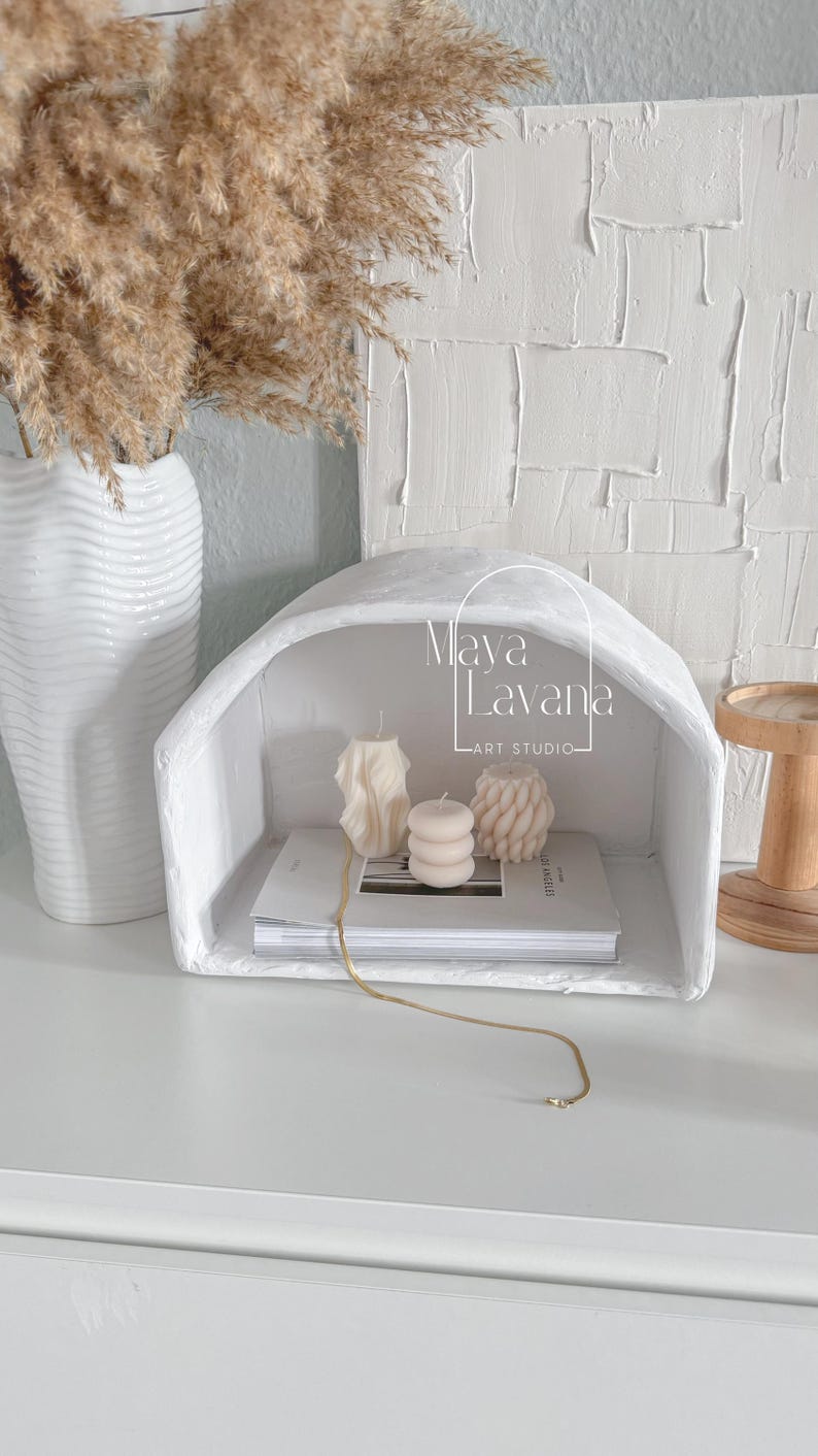 May include: White ceramic arched shelf with three candles and a stack of books. The shelf has the text "Maya Lavana - ART STUDIO" on the front.