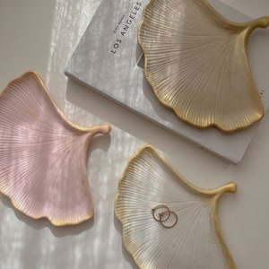 Könnte beinhalten: Drei blattförmige Dekotabletts in Rosa, Creme und Weiß mit goldenen Rändern. Zwei goldene Ringe liegen auf einem Tablett. Die Tabletts befinden sich auf einer weißen Oberfläche, im Hintergrund ein Buch.