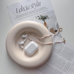 Puede incluir: Auriculares inalámbricos blancos y una funda de carga blanca están en un cuenco de cerámica blanco. Un collar de cuentas rosa y blanco está drapeado sobre el cuenco y una cadena dorada con un dije dorado cuelga del lado del cuenco.