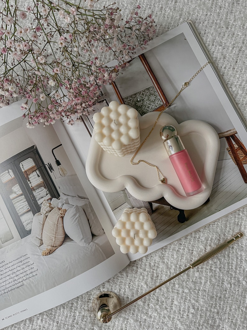 May include: A white ceramic tray with a cloud-like shape, featuring two white bubble candles and a pink lip balm tube. The tray is on a white surface with a gold chain necklace and a gold metal decorative stand.