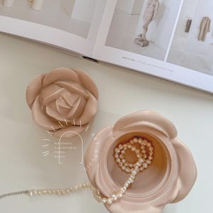May include: A light pink ceramic rose-shaped trinket box with a lid. The box is open and contains a strand of white pearls. The box is sitting on a white surface. The text "Aesthetic Sustainable Home Decor" is visible on the side of the box.