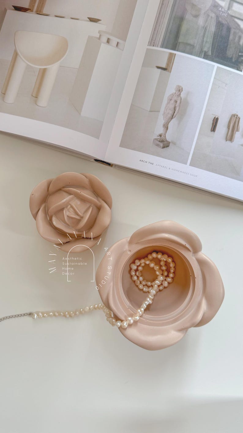 May include: A light pink ceramic rose-shaped trinket box with a lid. The box is open and contains a strand of white pearls. The box is sitting on a white surface. The text "Aesthetic Sustainable Home Decor" is visible on the side of the box.