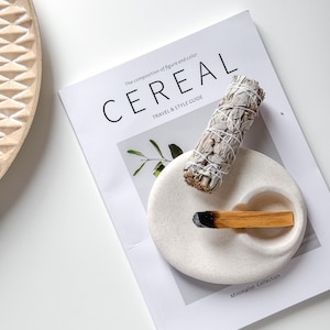 Könnte beinhalten: Weiße Keramik-Schale mit zwei Vertiefungen, eine mit einem weißen Salbei-Reinigungsstab und die andere mit einem Holzstab aus Palo Santo-Räucherwerk. Die Schale liegt auf einem Magazin mit dem Titel "Cereal" mit dem Text "The composition of figure and color" und "Travel & Style Guide".