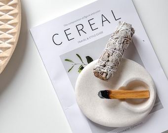 May include: White ceramic dish with two indentations, one holding a white sage smudge stick and the other holding a wooden stick of Palo Santo incense. The dish is on top of a magazine titled "Cereal" with the text "The composition of figure and color" and "Travel & Style Guide".