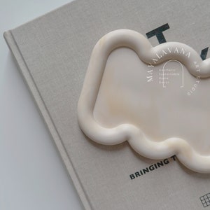 May include: A white ceramic tray with a wavy edge. The tray is on a beige book with the text "MAYALAVANA ART STUDIO" and "BRINGING THE SOHO" printed on it.