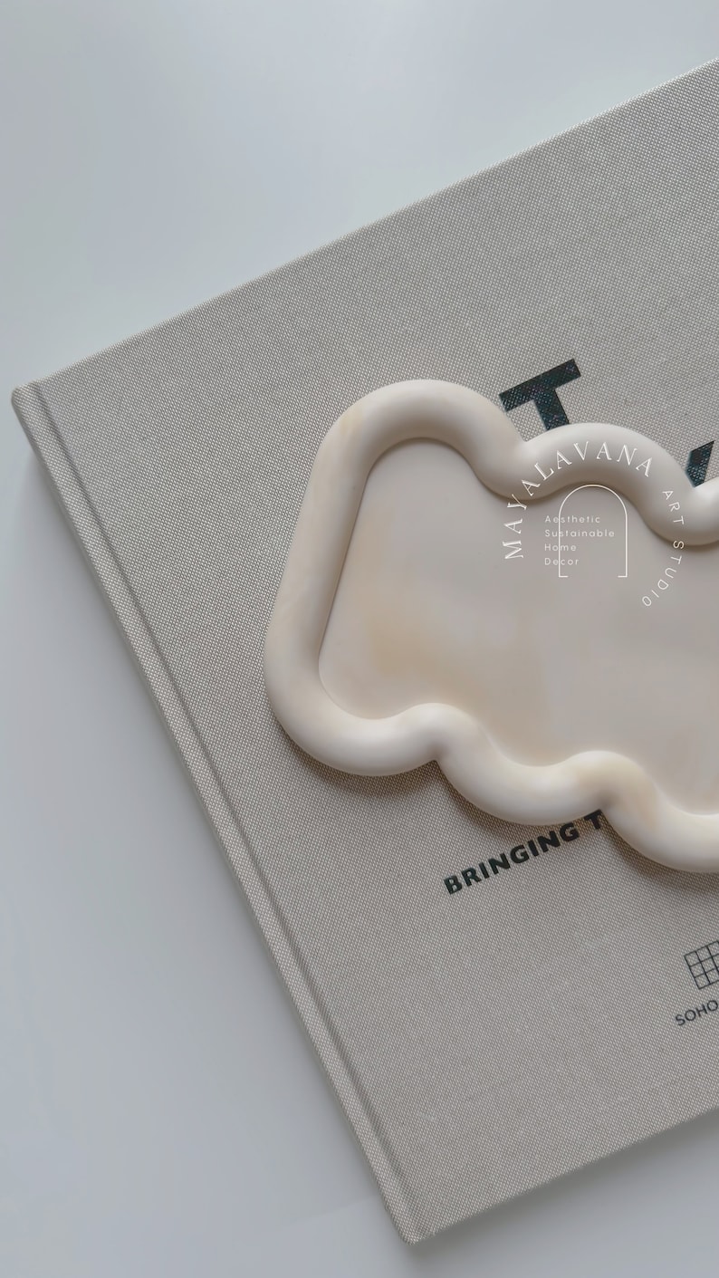 May include: A white ceramic tray with a wavy edge. The tray is on a beige book with the text "MAYALAVANA ART STUDIO" and "BRINGING THE SOHO" printed on it.