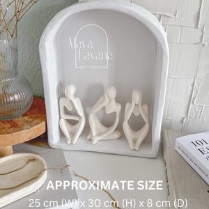May include: White ceramic sculpture of three figures sitting with their heads in their hands. The sculpture is displayed in a white arched frame. The frame is 25 cm wide, 30 cm high, and 8 cm deep. The text "Maya Lavana Art Studio" is visible on the frame.