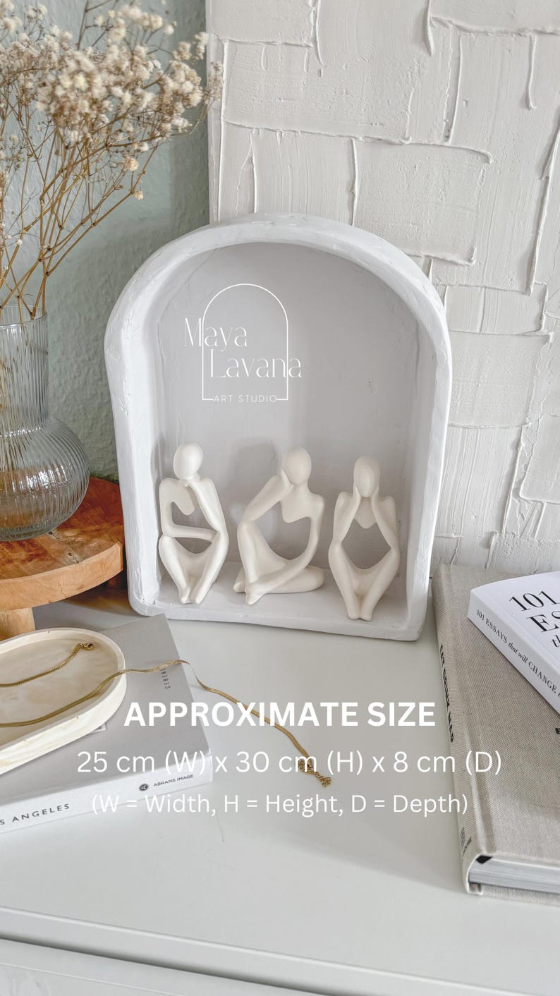 May include: White ceramic sculpture of three figures sitting with their heads in their hands. The sculpture is displayed in a white arched frame. The frame is 25 cm wide, 30 cm high, and 8 cm deep. The text "Maya Lavana Art Studio" is visible on the frame.