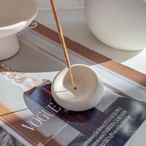 Könnte beinhalten: Ein weißer, runder, organisch geformter Kerzenhalter aus Keramik. Ein brauner Räucherstäbchen brennt im Halter. Der Halter steht auf einem Magazin mit dem Wort "VOGUE" auf dem Cover.
