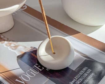May include: A white ceramic incense holder with a round, organic shape. A brown incense stick is burning in the holder. The holder is sitting on a magazine with the word "VOGUE" printed on the cover.