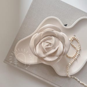 May include: A white ceramic rose sculpture sits on a white ceramic tray with a pearl necklace. The tray is shaped like a bird. The background is a light gray fabric.