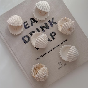 Może przedstawiać: Białe muszelki jako uchwyty na pierścionki ułożone na książce z napisem "EAT DRINK NAP" i "BRINGING THE HOUSE HOME". Niektóre muszle trzymają złote pierścionki. Muszle mają teksturowaną powierzchnię.