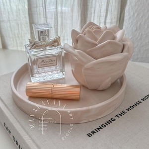 May include: A white ceramic tray with a rose-shaped decorative object and a bottle of perfume labeled "Miss Dior Blooming Bouquet". A gold lipstick is also on the tray. The tray is sitting on a book with the text "Bringing the House Home" printed on the cover.