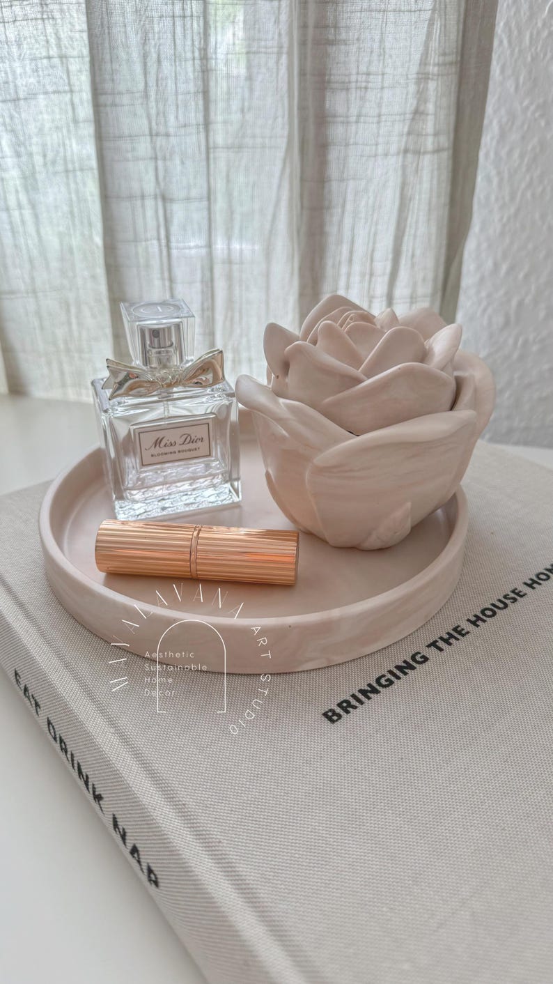 May include: A white ceramic tray with a rose-shaped decorative object and a bottle of perfume labeled "Miss Dior Blooming Bouquet". A gold lipstick is also on the tray. The tray is sitting on a book with the text "Bringing the House Home" printed on the cover.