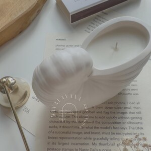May include: A white ceramic candle holder shaped like a seashell with a white candle inside. The candle holder is sitting on a book with the text "Aesthetic Sustainable Home Decor" and "Artist" visible. There are also some white matchsticks and a box of matches with the text "Be Salt and Light" on it.