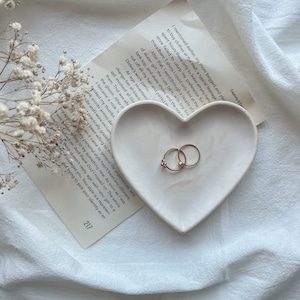 Puede incluir: Un plato blanco en forma de corazón con dos anillos de oro y un anillo de piedra azul sobre un fondo de tela blanca. El plato está sobre un trozo de papel con texto que dice "Creatividad en Ep Mencioné antes que mi madre rogó y suplicó para comprar ropa en el centro comercial, un típico adolescente..."