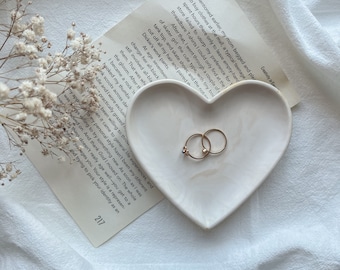 May include: A white heart-shaped dish with two gold rings and a blue gemstone ring on a white fabric background. The dish is sitting on a piece of paper with text that reads "Creativity in Ep I mentioned earlier, my mom begged and pleaded to buy clothes at the mall, a typical teenage..."