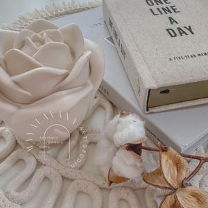 May include: A white ceramic rose sits on a white woven rug with a book titled "One Line a Day: A Five-Year Memory Journal" and a cotton boll.