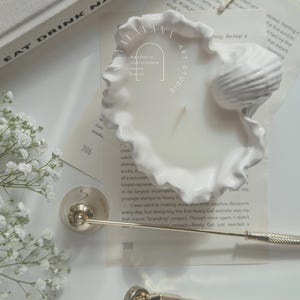 May include: White ceramic candle holder shaped like a seashell with a white candle inside. The candle holder is sitting on a white surface with a book and a pair of scissors.