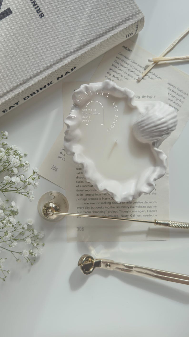May include: White ceramic candle holder shaped like a seashell with a white candle inside. The candle holder is sitting on a white surface with a book and a pair of scissors.