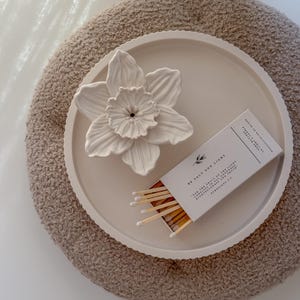 Può includere: Un fiore di narciso in ceramica bianca e una scatola di fiammiferi su un vassoio rotondo bianco sporco. Il vassoio è su un pouf circolare beige strutturato. La scatola di fiammiferi recita "BE SALT AND LIGHT".