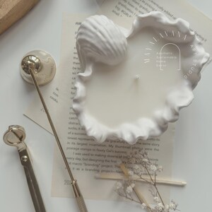 May include: A white ceramic seashell candle holder with a white candle inside. The candle holder is on a white surface with a gold candle snuffer and a box of matches. The text "Aesthetic Sustainable Home Decor" is visible on the candle holder.