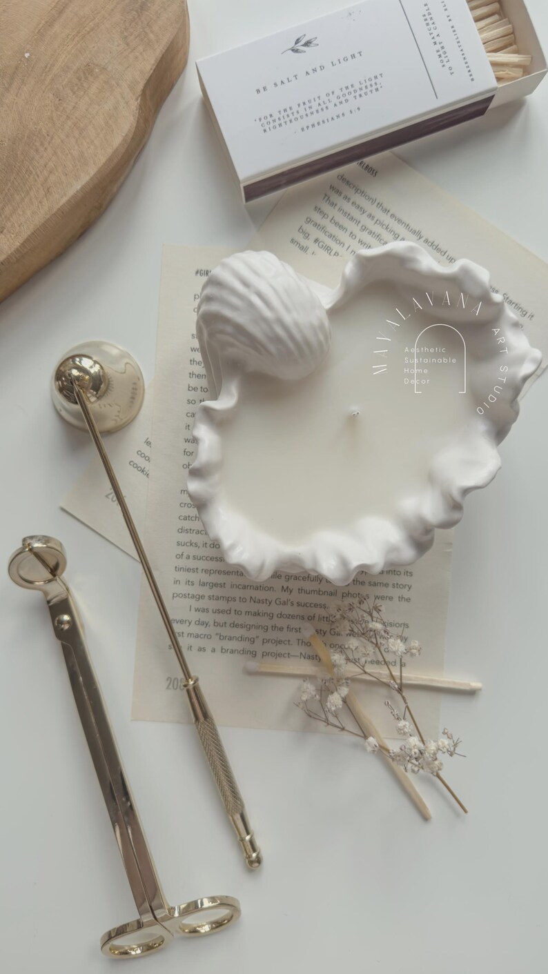 May include: A white ceramic seashell candle holder with a white candle inside. The candle holder is on a white surface with a gold candle snuffer and a box of matches. The text "Aesthetic Sustainable Home Decor" is visible on the candle holder.