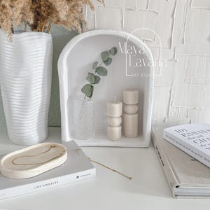 May include: White ceramic shelf with an arched top, featuring two white candles with a textured design, a clear glass vase with a sprig of eucalyptus, and the text "Maya LaVarie Art Studio".