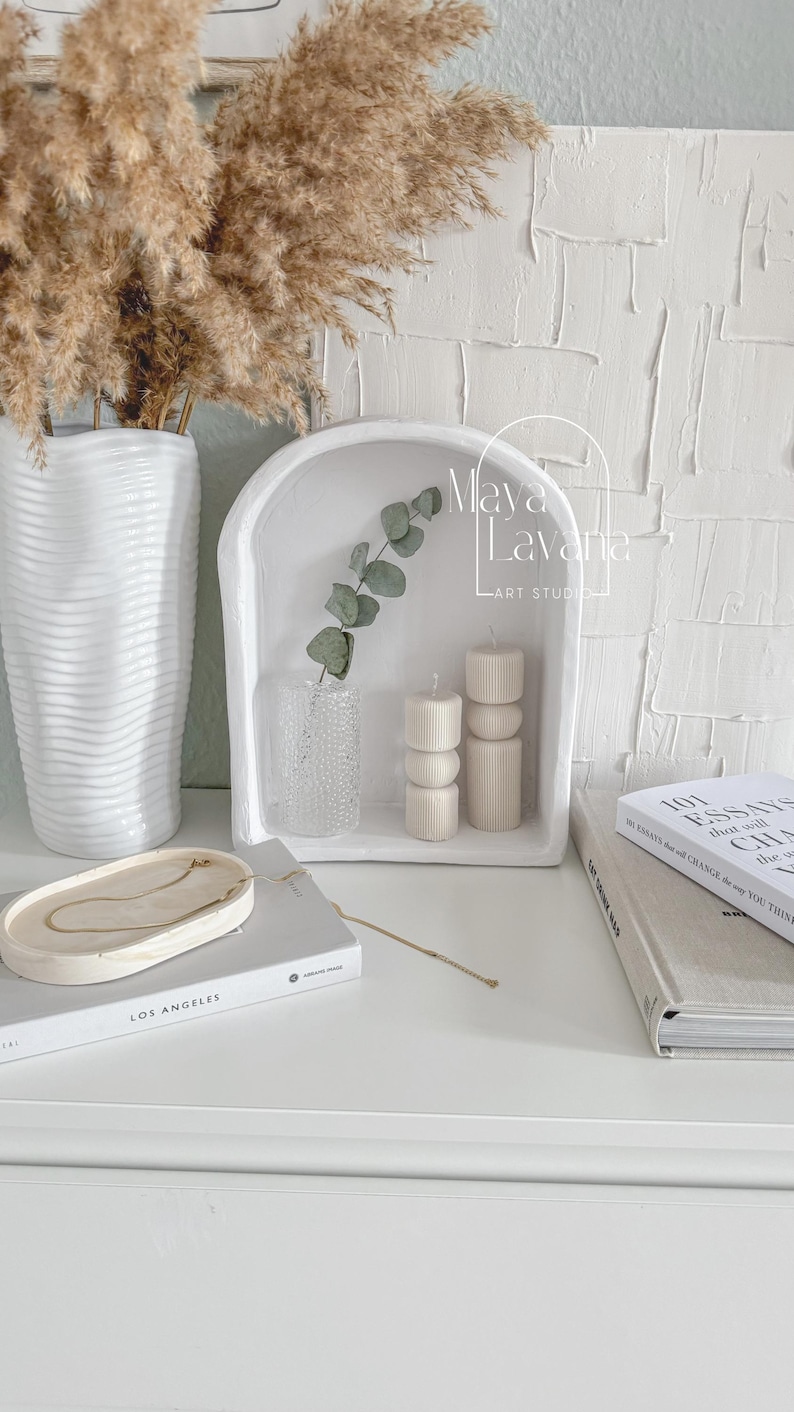 May include: White ceramic shelf with an arched top, featuring two white candles with a textured design, a clear glass vase with a sprig of eucalyptus, and the text "Maya LaVarie Art Studio".