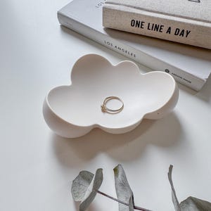 Puede incluir: Plato para anillos de cerámica blanca en forma de nube con un anillo de oro en su interior. El plato está sobre una superficie blanca con una rama de hojas de eucalipto.