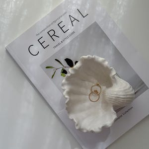 以下が含まれることがあります： 白い貝殻型のジュエリー皿に2つのゴールドリングが置かれています。「CEREAL」という旅行とスタイルのガイド雑誌の上にあります。自然光が柔らかい影を作り出しています。