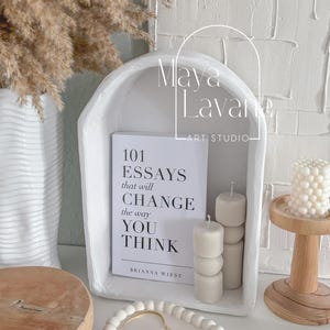 May include: White ceramic arch with a book titled "101 Essays that will CHANGE the way YOU THINK" by Brianna Wiest. The arch is decorated with white candles and a small wooden tray with white beads.