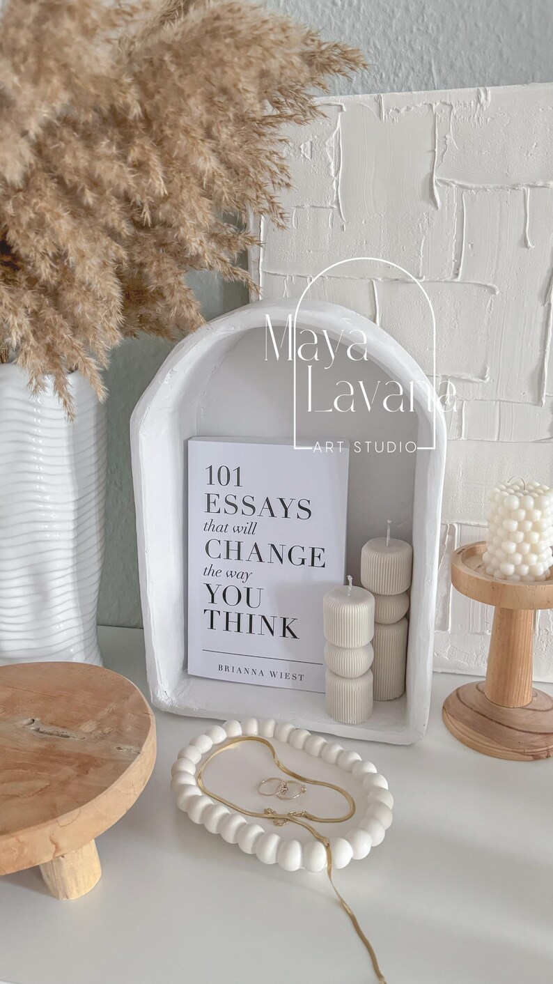 May include: White ceramic arch with a book titled "101 Essays that will CHANGE the way YOU THINK" by Brianna Wiest. The arch is decorated with white candles and a small wooden tray with white beads.